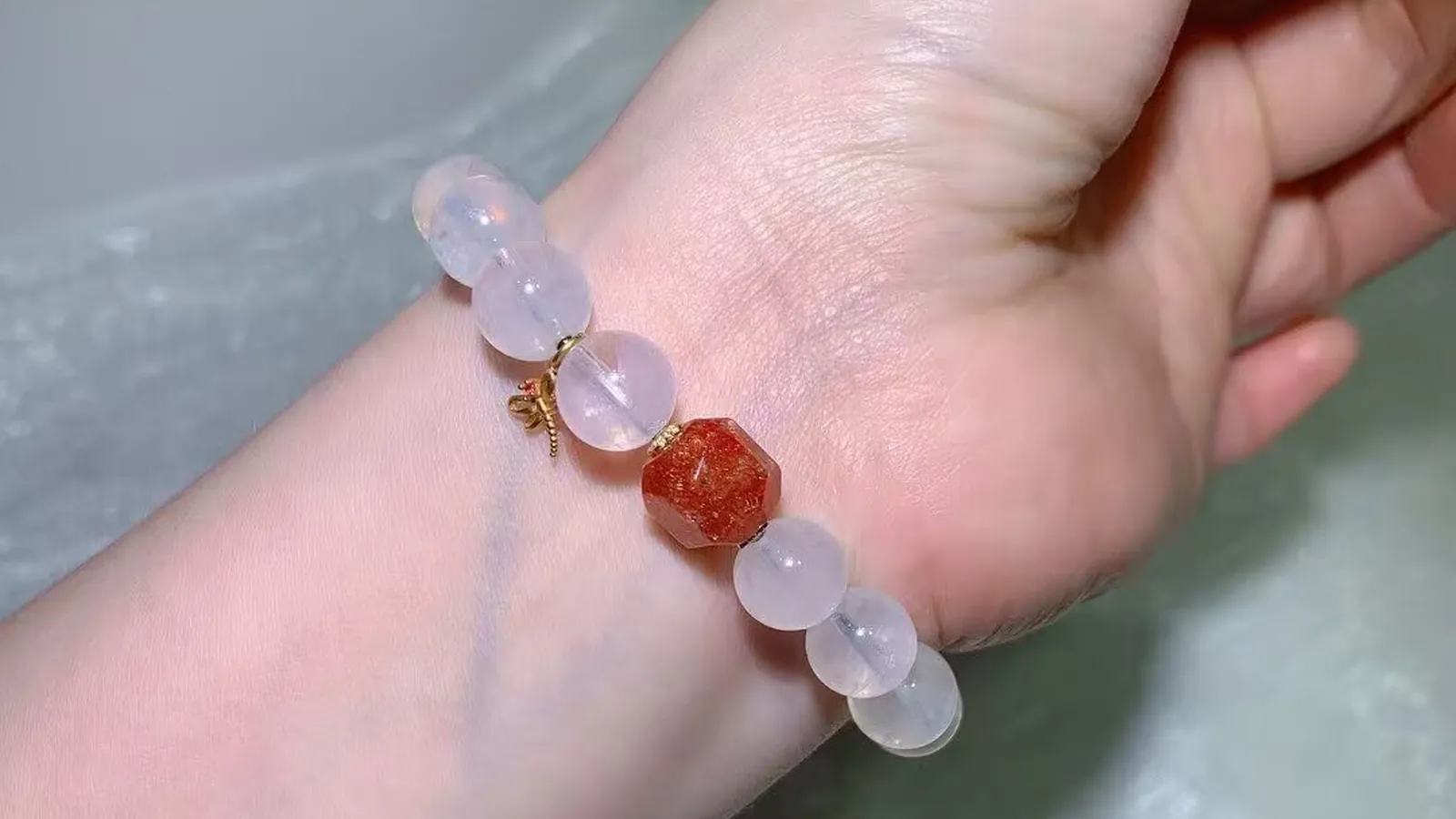 Blue needle quartz bracelet on a wrist: icy milky-white beads with a faceted coral-orange bead and a small vintage-style charm