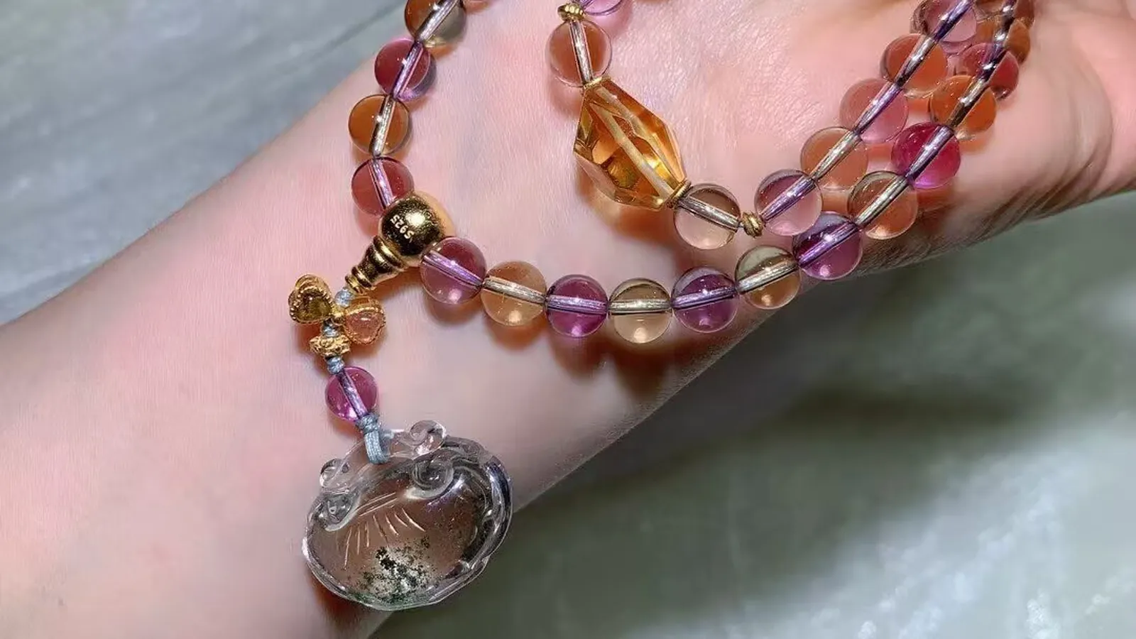 An ametrine 108-bead mala necklace wrapped around the wrist, purple and honey-yellow beads with a vintage-style guru bead and a carved green phantom quartz charm